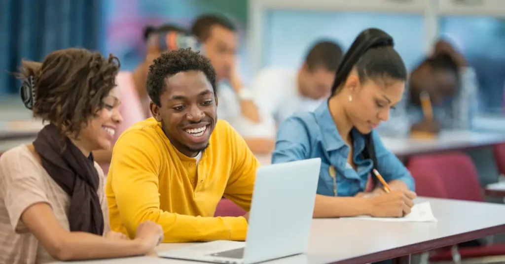 Three college students working on an assignment in a classroom. Two are looking at a laptop together and one is writing on a piece of paper.