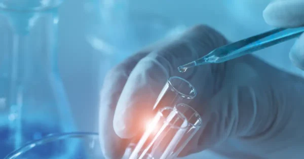 Image of a hand with a latex glove holding a glass test tube and using a pipette to drop liquid into the test tube.