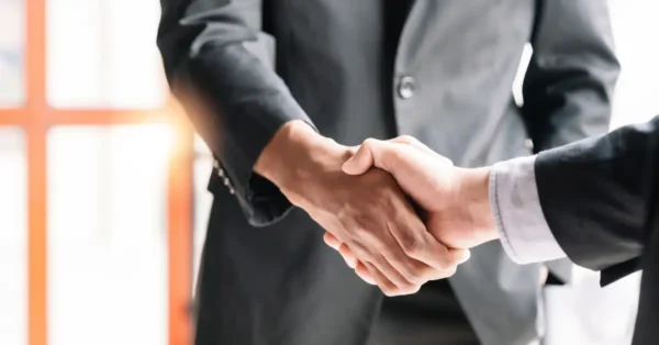 Two individuals wearing dark suits, shaking hands to represent a merger and acquisition business agreement.