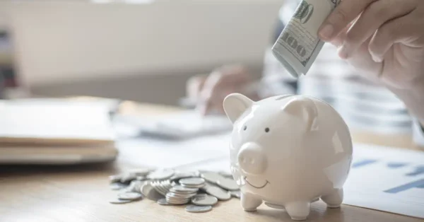 A white ceramic piggy bank on a wooden table, with coins piled to the side and a $100 bill being placed in the bank slot. This image represents the concept of increasing Health Savings Account (HSA) contributions. The piggy bank symbolizes savings, and the $100 bill suggests an increase in funds allocated to an HSA.