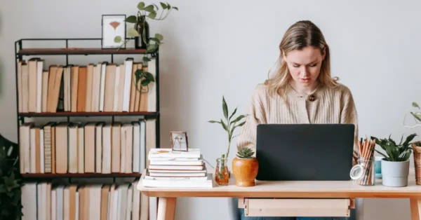 A female with blonde hair, sitting at a desk and looking at her laptop, meant to represent a first-year audit associate at a CPA firm. 